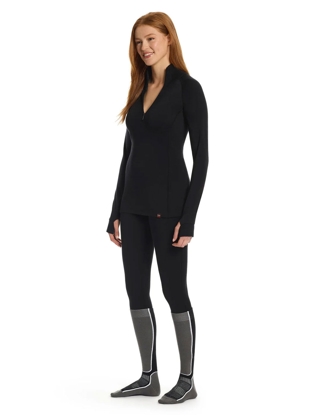 A woman modeling a black long-sleeve V-neck base layer top and matching black leggings, paired with gray and black leg warmers, against a plain white background.