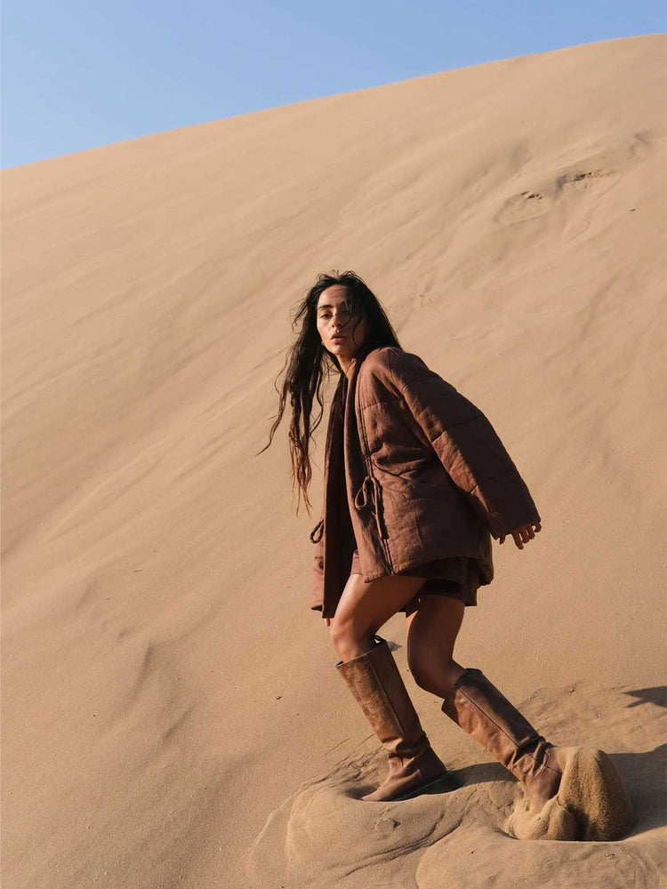 A woman posing on a sandy desert dune wearing a brown textured jacket, brown shorts, and matching brown boots under a clear blue sky.