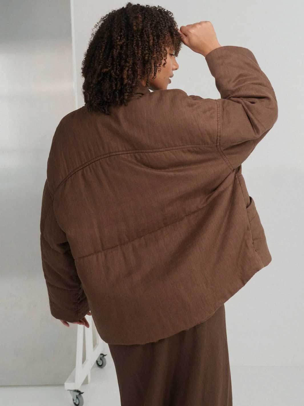 A person shown from the back wearing a brown quilted jacket and matching brown skirt in a studio setting.