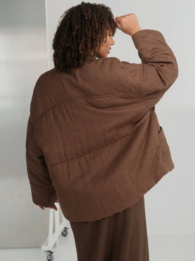 A person shown from the back wearing a brown quilted jacket and matching brown skirt in a studio setting.