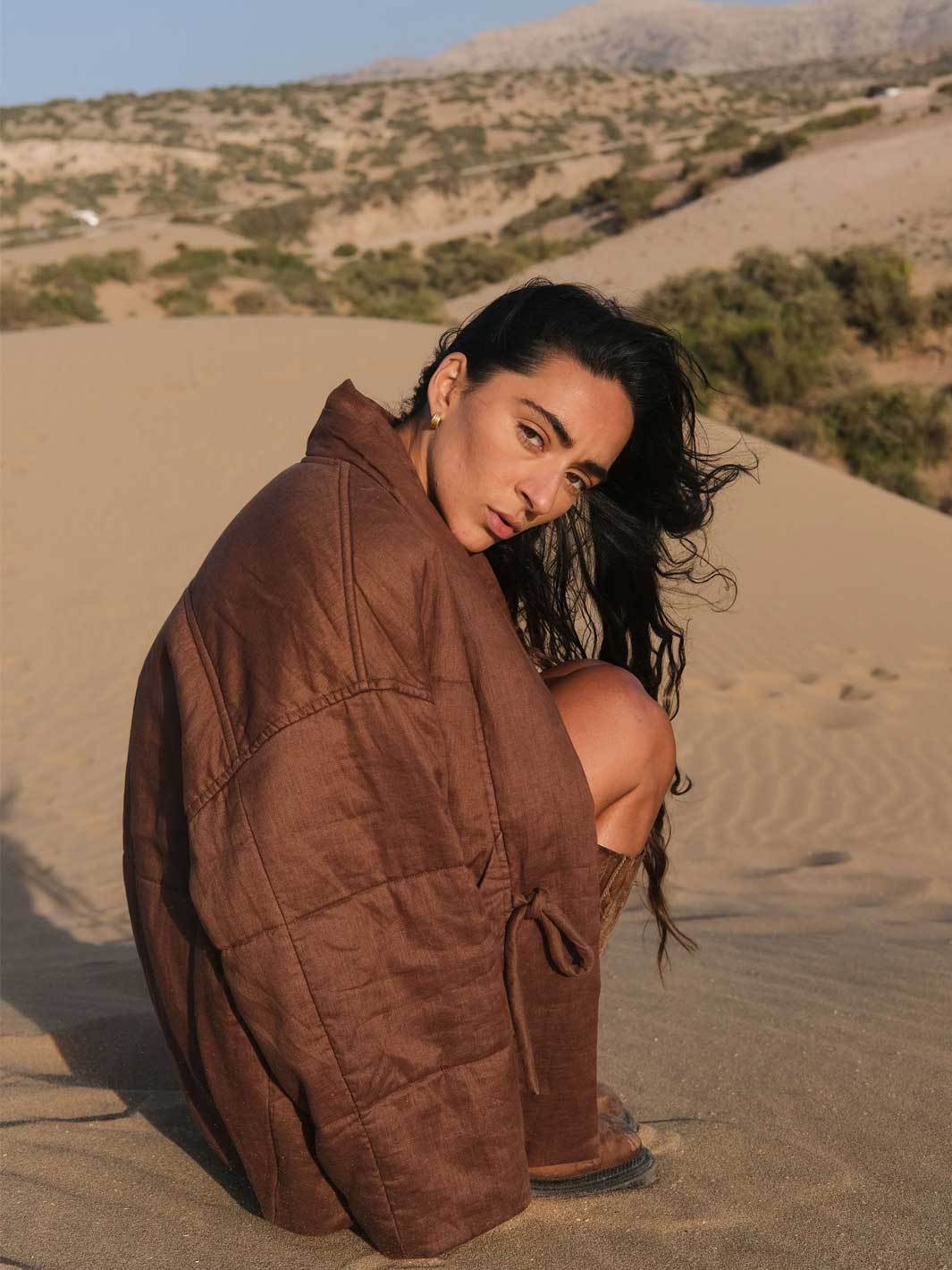 A woman crouches on a sandy desert dune wearing a brown quilted wrap jacket, with desert hills and sparse shrubs in the background.