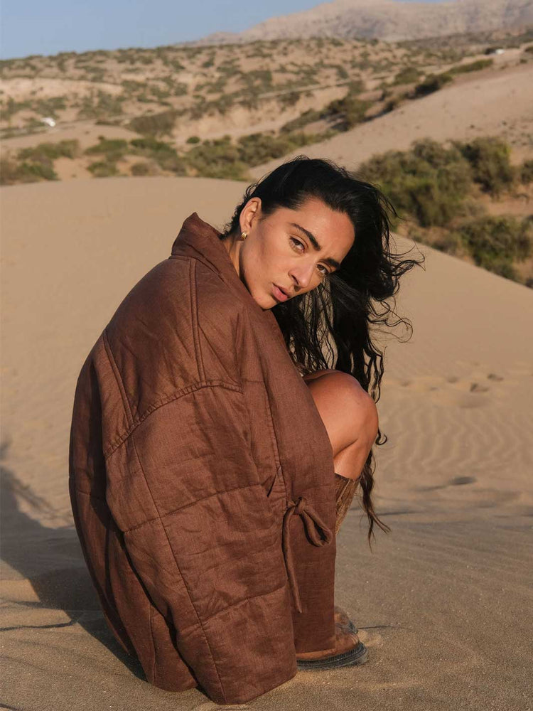 A woman crouches on a sandy desert dune wearing a brown quilted wrap jacket, with desert hills and sparse shrubs in the background.