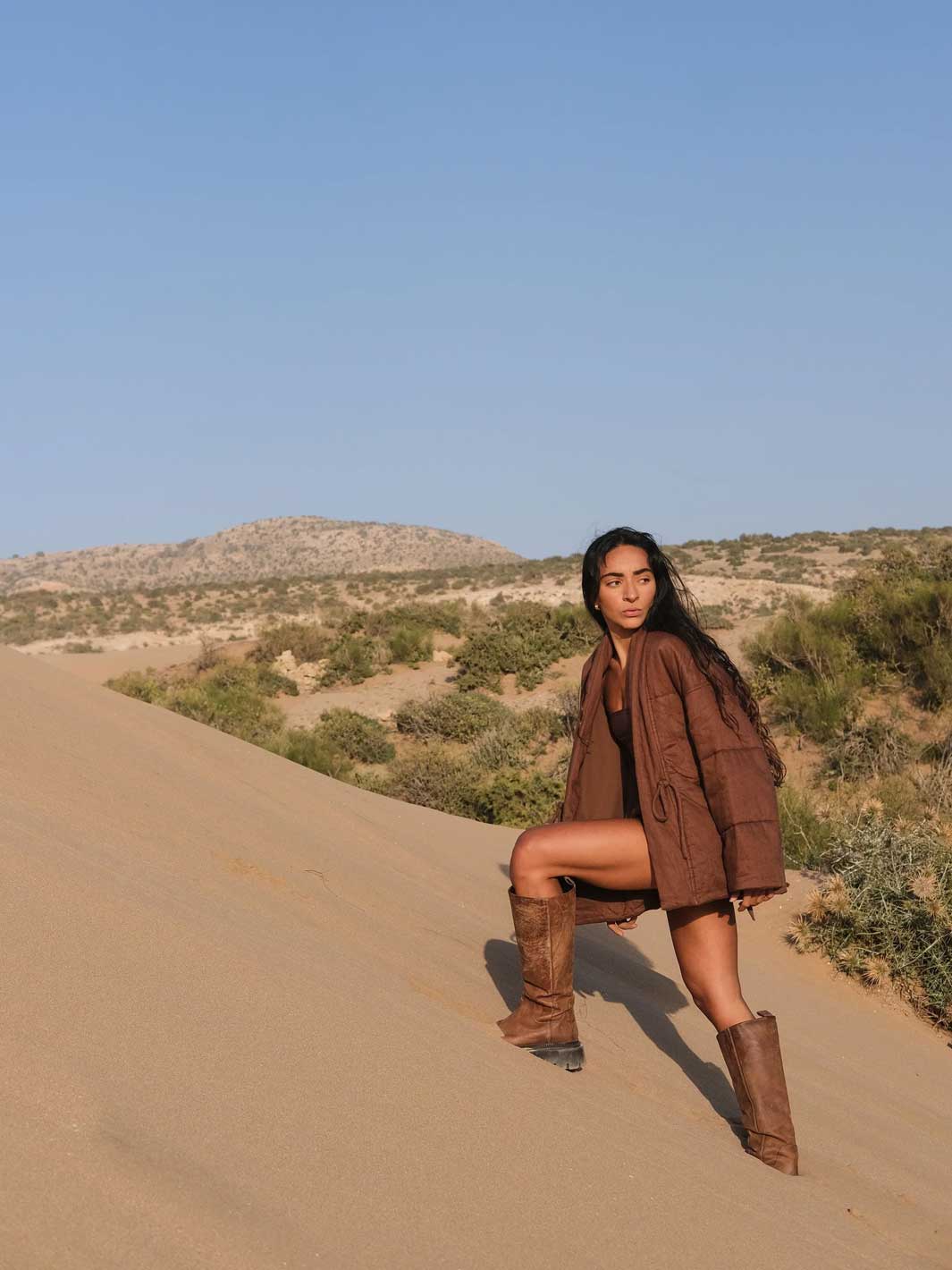 A woman posing on a desert sand dune wearing a brown padded linen jacket, black top, and matching brown boots, with a desert landscape and blue sky in the background.
