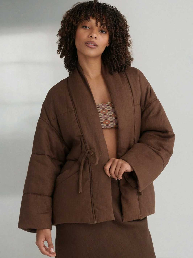 A model with curly dark hair wearing a brown linen puffer jacket with a shawl collar and drawstring, styled over a patterned bikini top and matching brown bottoms, set against a neutral light grey background.