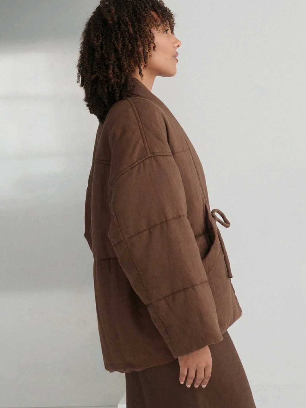 A model shown in profile wearing a brown quilted jacket with a matching belt and brown skirt against a plain light grey background.
