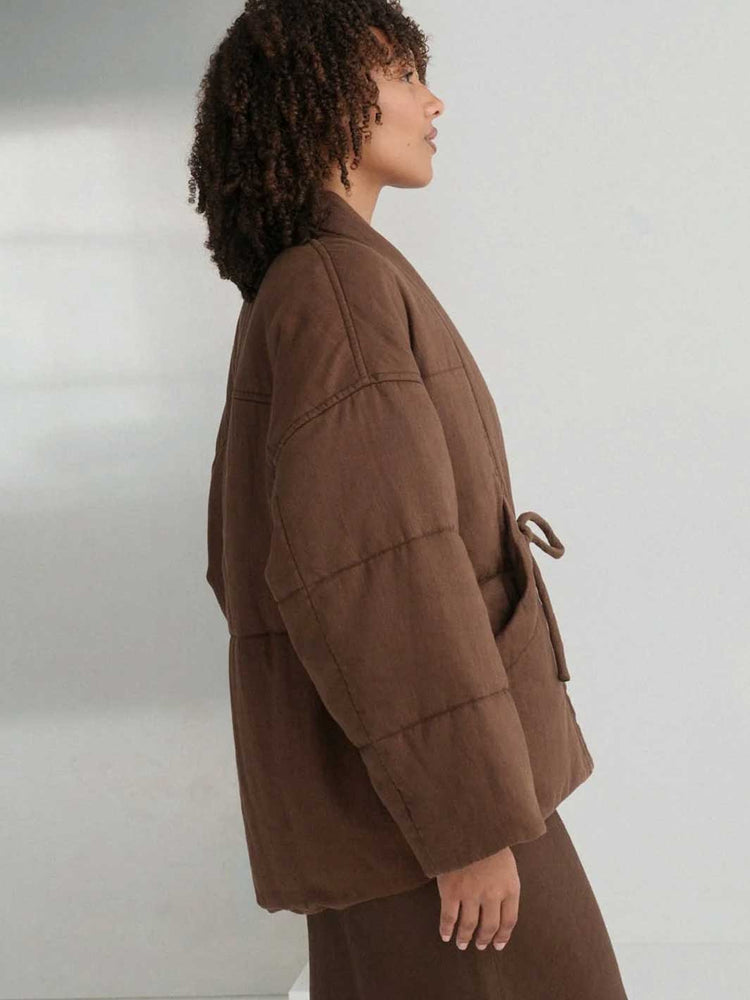 A model shown in profile wearing a brown quilted jacket with a matching belt and brown skirt against a plain light grey background.