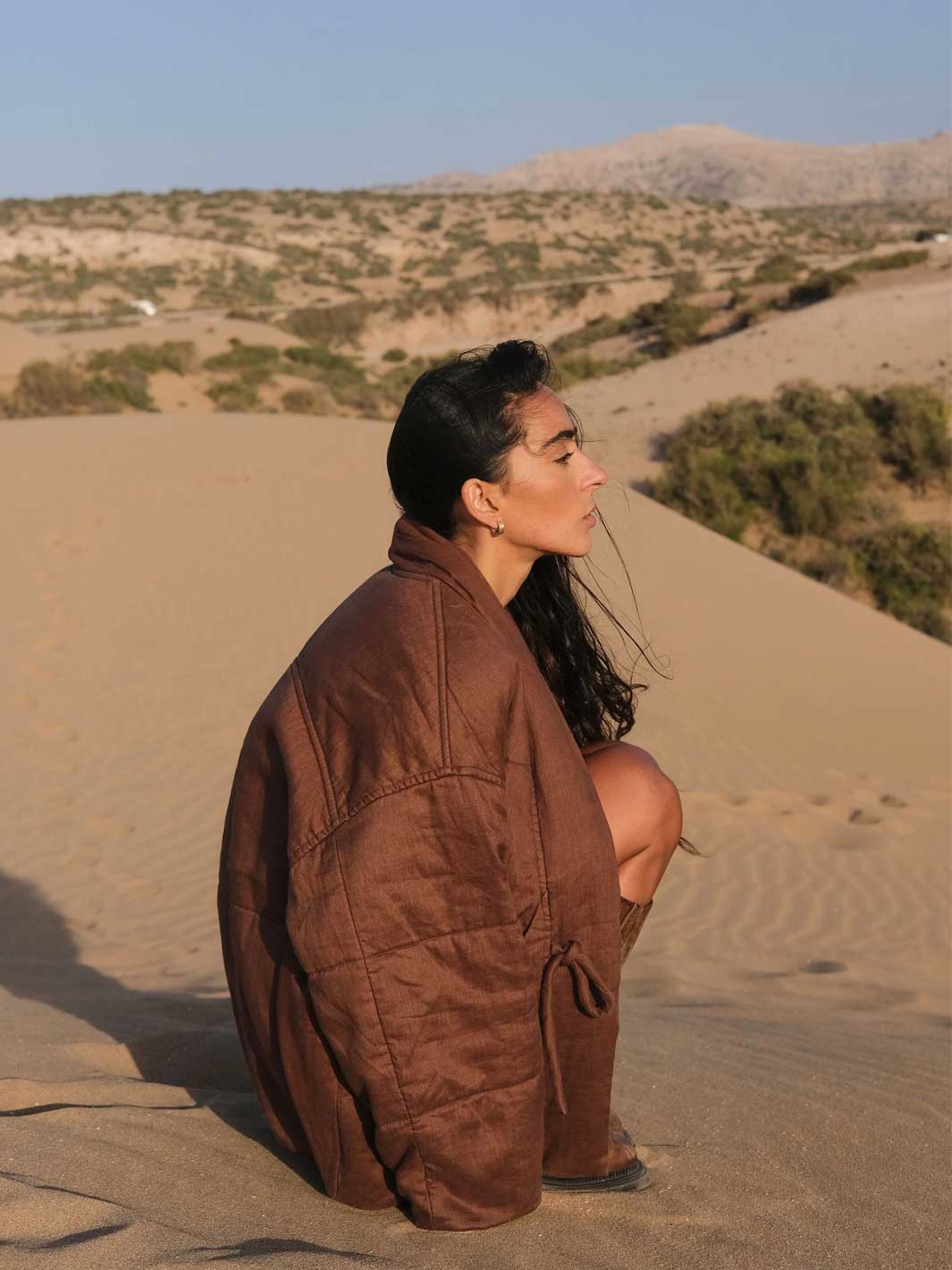 A woman crouches on a sandy dune in a desert landscape, wearing a loose brown padded jacket. The background shows sand, sparse shrubs, and distant hills under a clear blue sky.