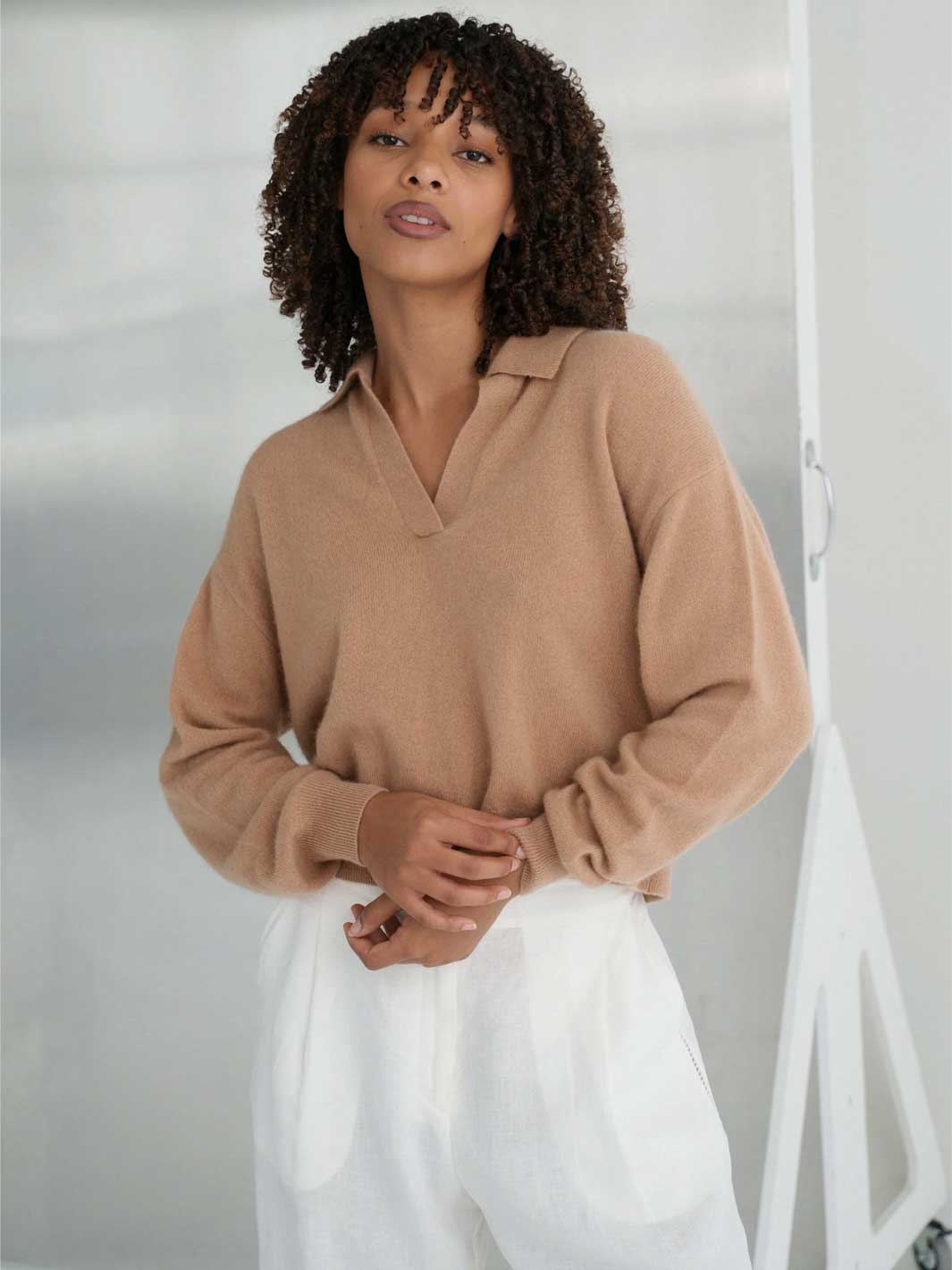A woman with curly dark hair wearing a camel-colored knit polo sweater and white trousers, standing against a light gray studio background.