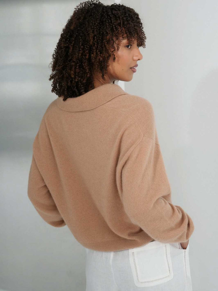 A woman with curly dark hair is shown from the back wearing a camel-colored knit sweater and white pants against a light gray background.