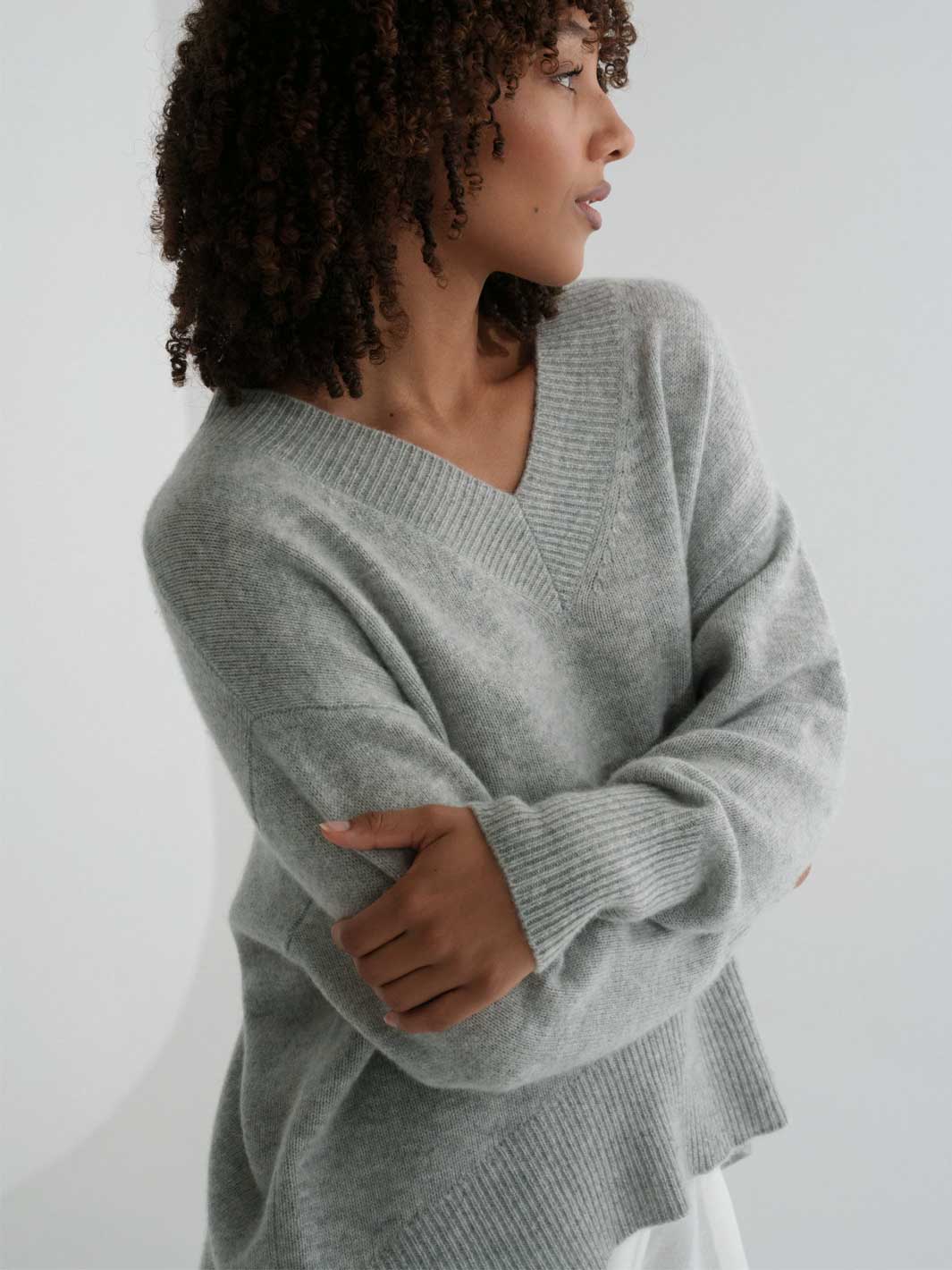 A woman wearing a light gray ribbed knit sweater with a V-neck and long sleeves, shown from the side against a plain light background.