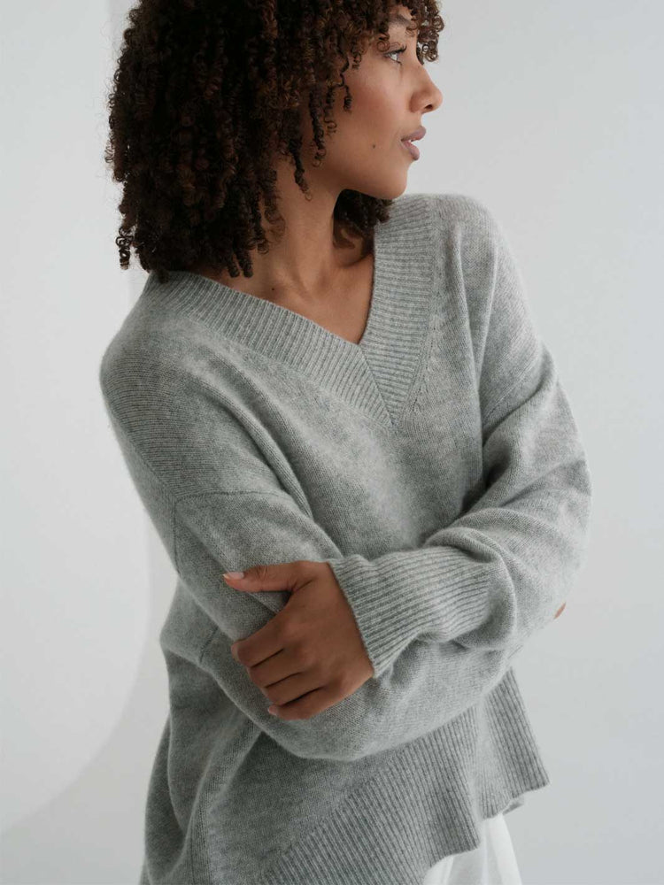 A woman wearing a light gray ribbed knit sweater with a V-neck and long sleeves, shown from the side against a plain light background.