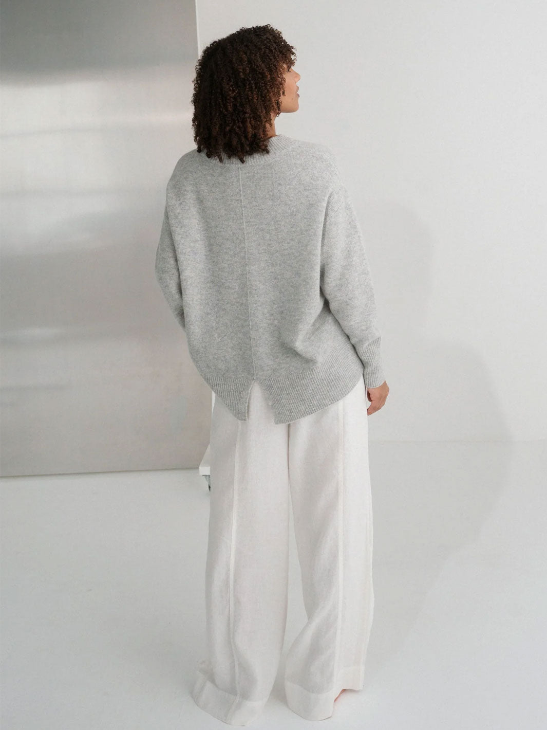 A model shown from the back wearing a light grey oversized knit sweater and white wide-leg trousers in a minimalist studio setting.