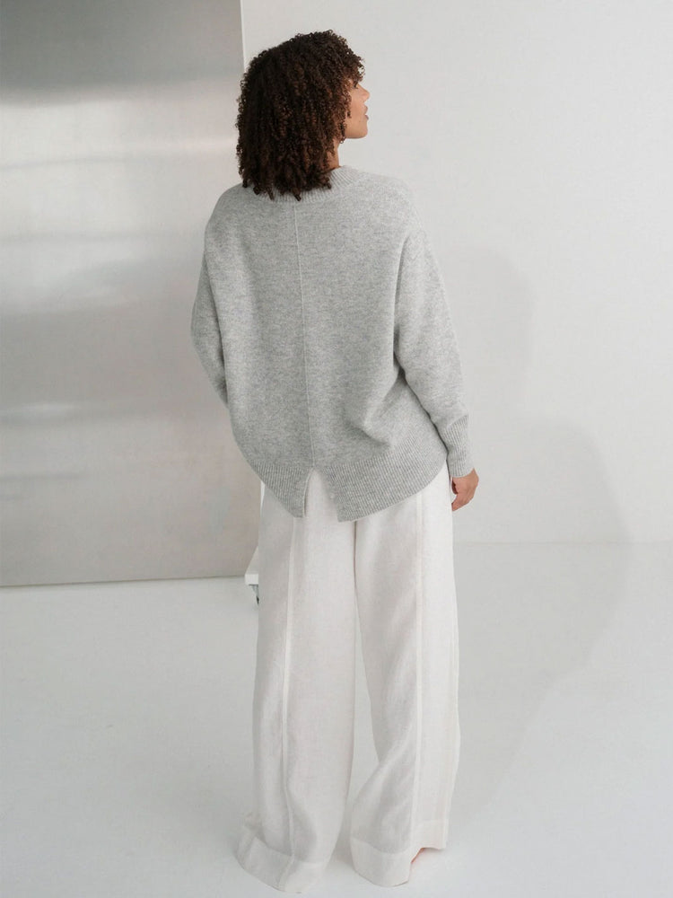 A model shown from the back wearing a light grey oversized knit sweater and white wide-leg trousers in a minimalist studio setting.
