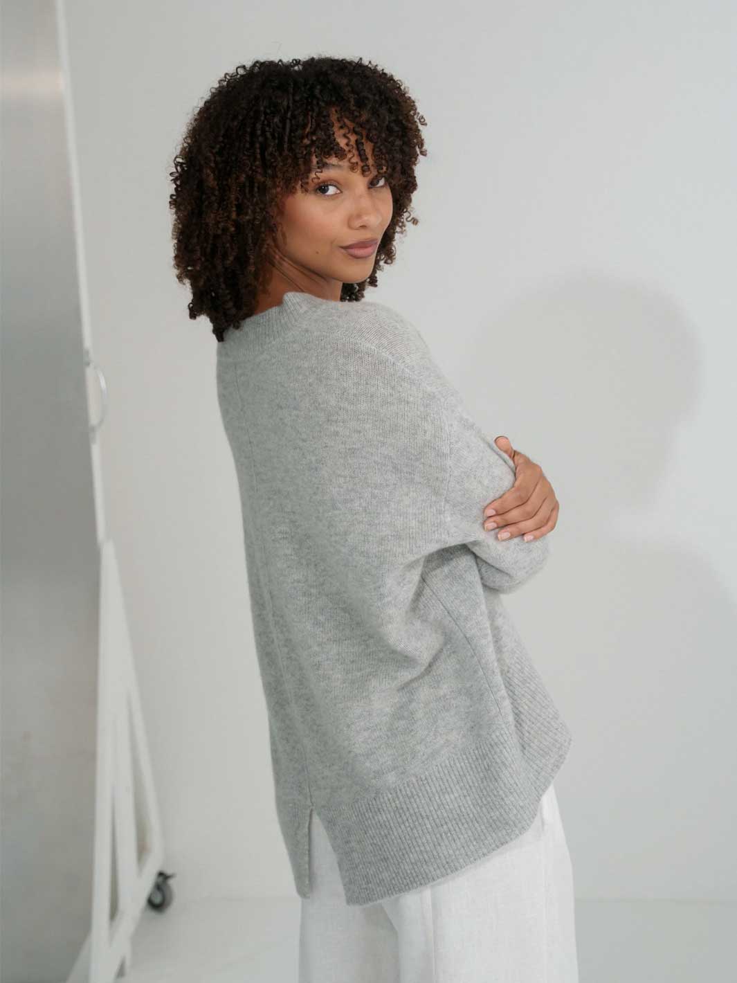 A woman with short curly dark hair is shown from the back wearing an oversized light gray knit sweater and white pants in a bright white studio setting.
