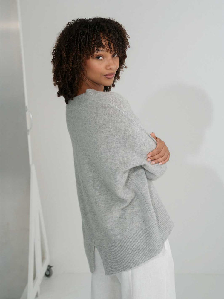 A woman with short curly dark hair is shown from the back wearing an oversized light gray knit sweater and white pants in a bright white studio setting.