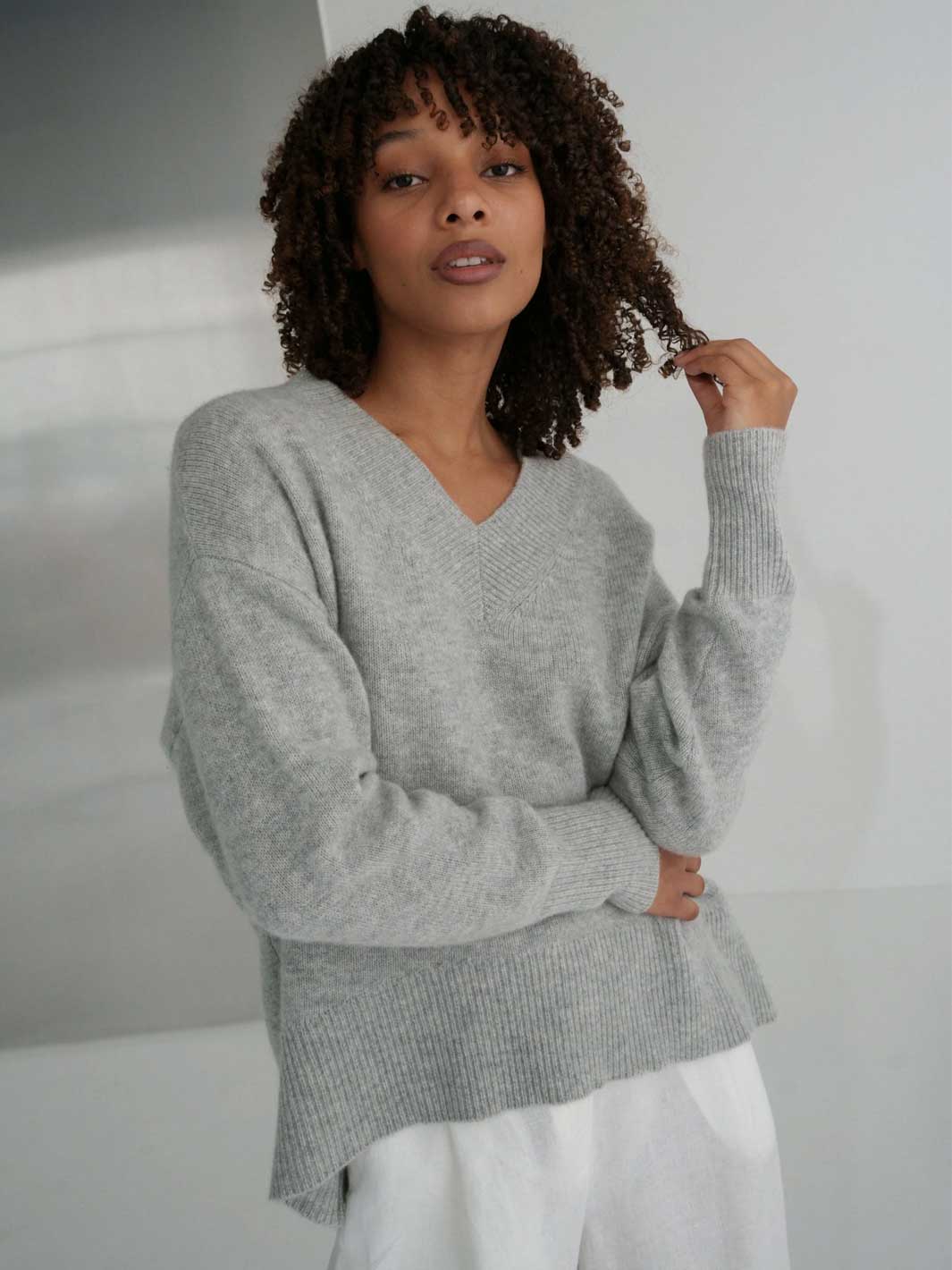 A woman with curly dark hair wearing a light grey heathered ribbed knit sweater with a V-neck and long sleeves, paired with white pants, standing against a light grey wall.