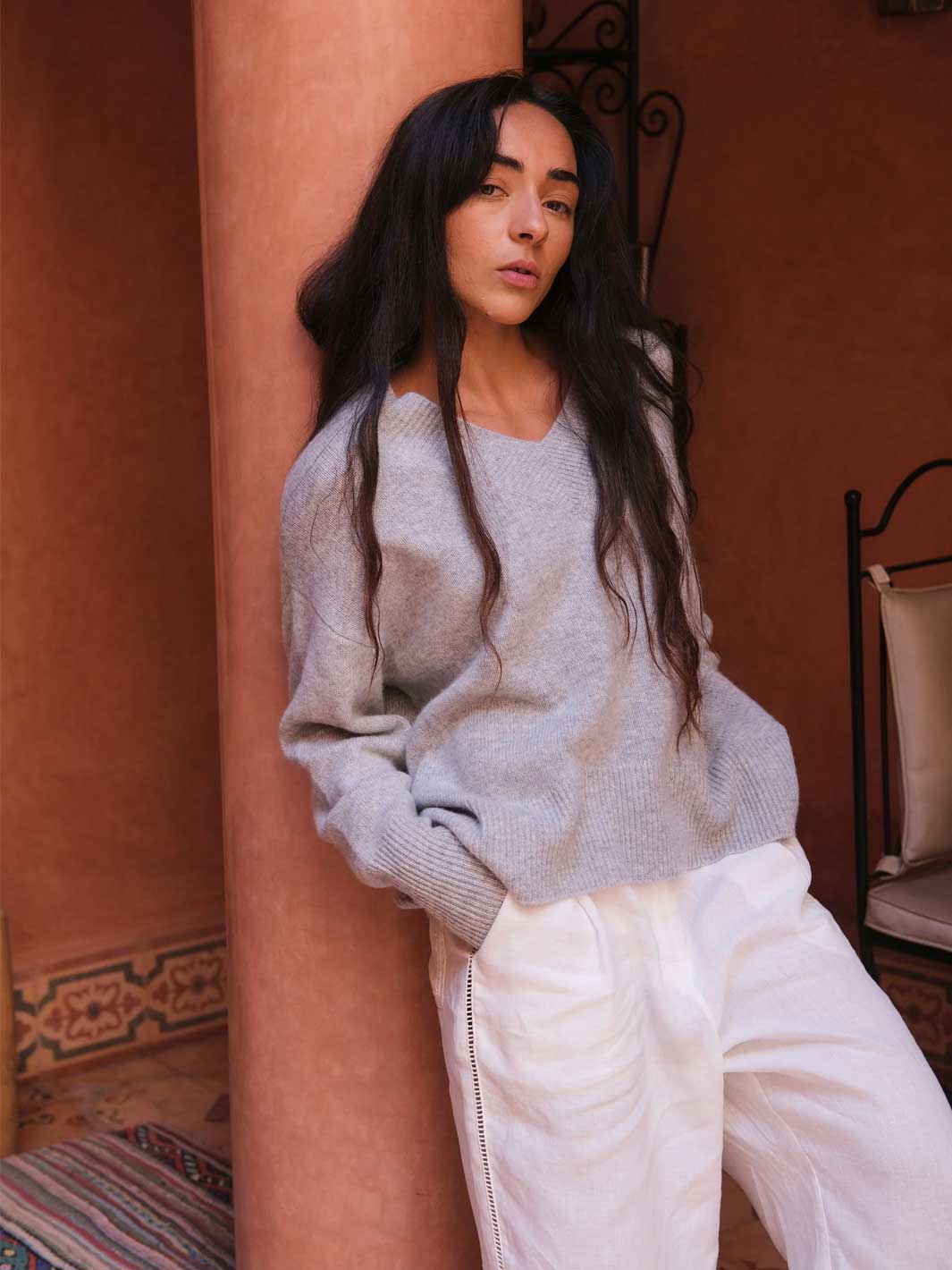 A woman with long dark hair leans against a terracotta-colored pillar, wearing a light grey knit sweater and white wide-leg pants in a warm-toned indoor setting.