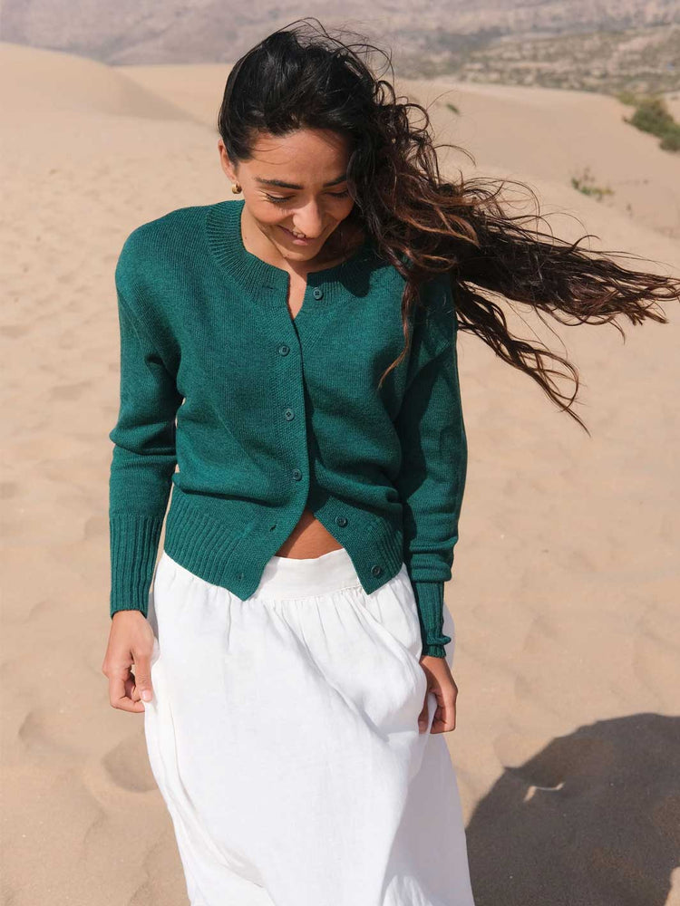 A woman standing on a sandy dune wearing a dark teal green button-up cardigan and a white skirt.