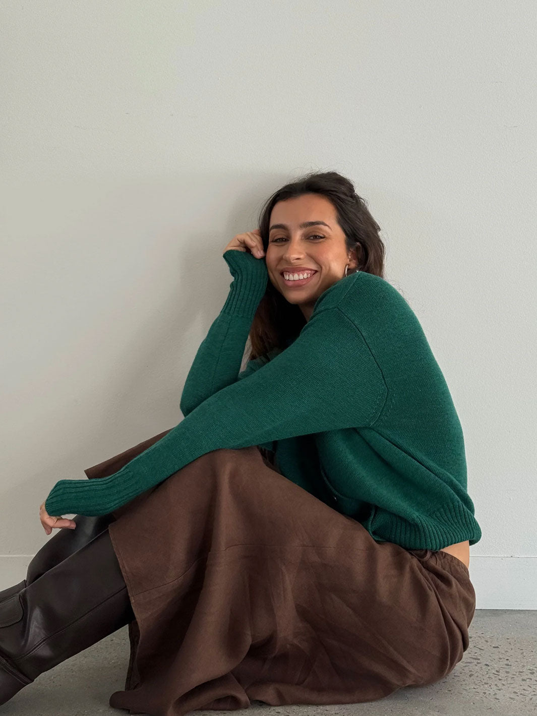 A woman sitting on a concrete floor against a plain light gray wall, wearing a dark green knit sweater, a brown skirt, and black boots.