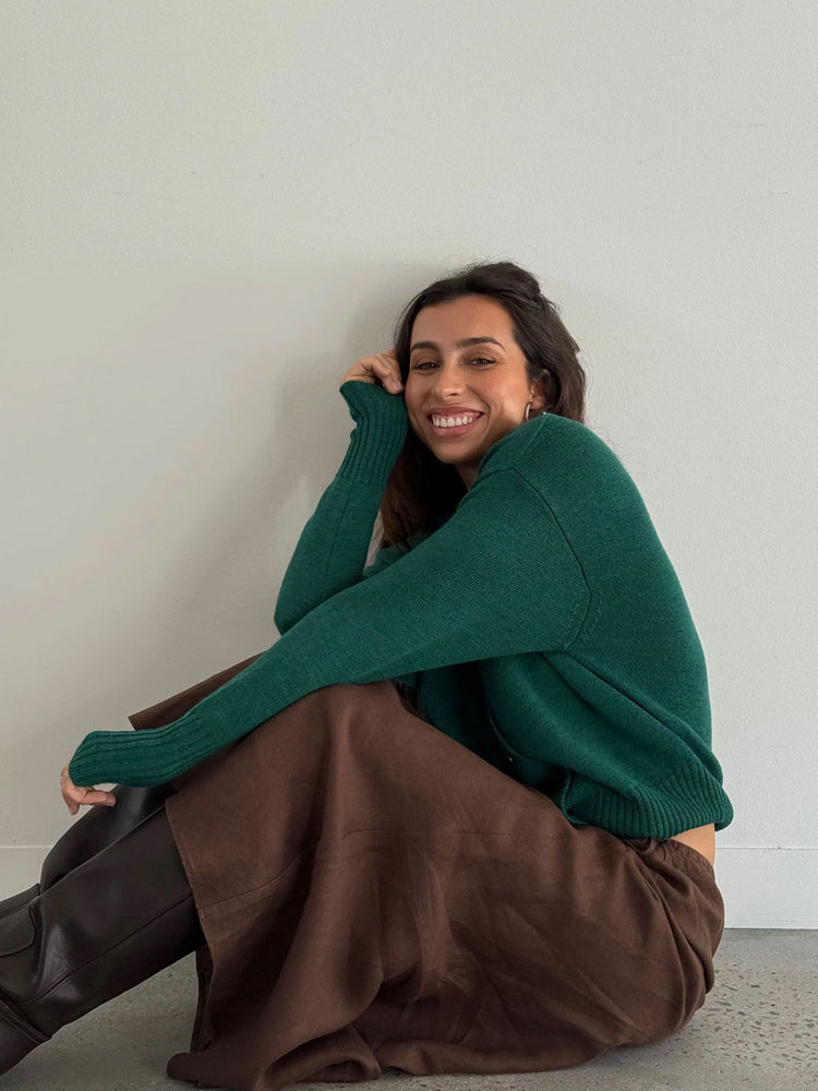 A woman sitting on a concrete floor against a plain light gray wall, wearing a dark green knit sweater, a brown skirt, and black boots.