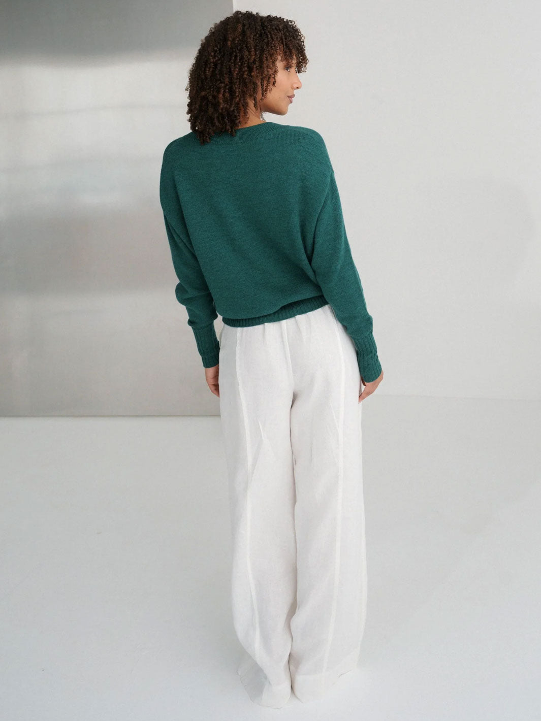 A woman shown from behind wearing a dark green long-sleeve knit sweater and white wide-leg trousers, standing against a minimalist light gray studio background.