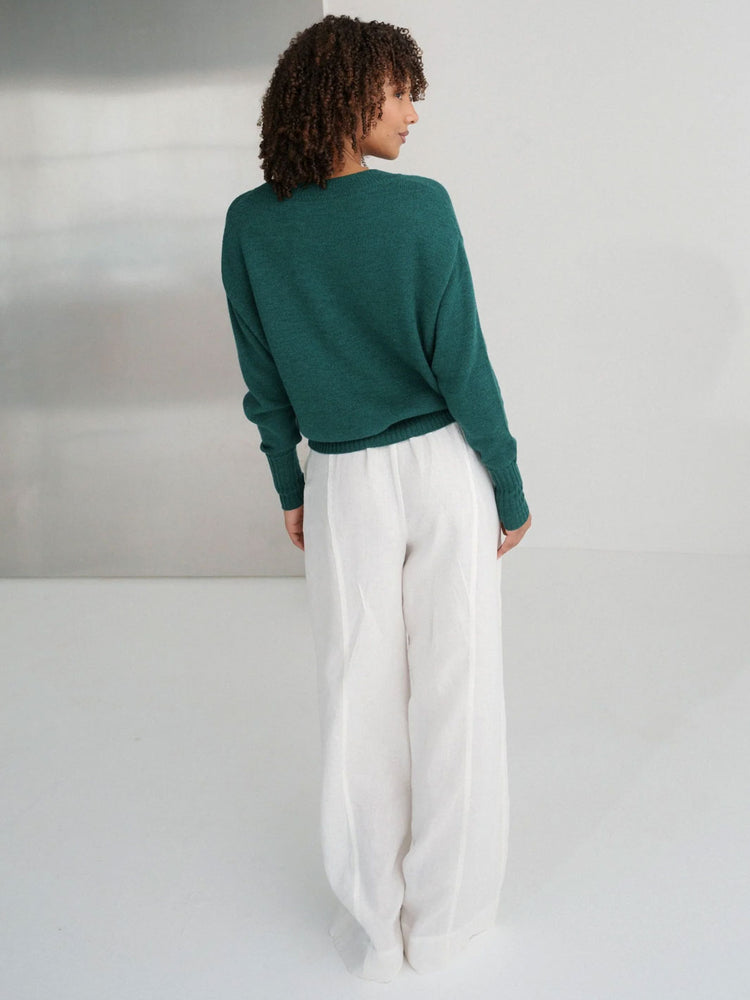 A woman shown from behind wearing a dark green long-sleeve knit sweater and white wide-leg trousers, standing against a minimalist light gray studio background.