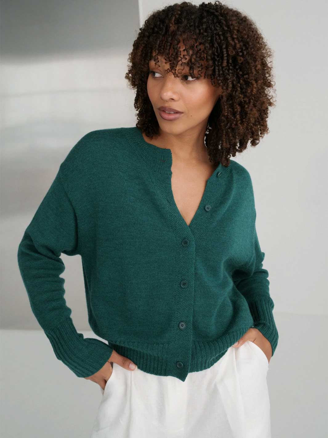 A woman with curly brown hair wearing a dark green button-up cardigan sweater and white trousers, standing against a light gray background.