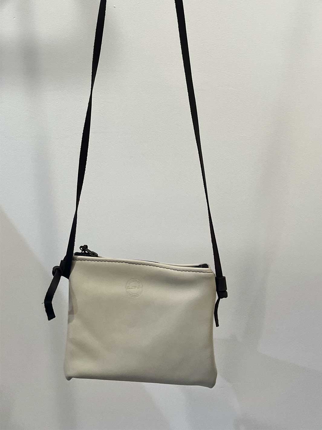 A small off-white leather crossbody bag with a dark brown shoulder strap, hanging against a plain white wall.
