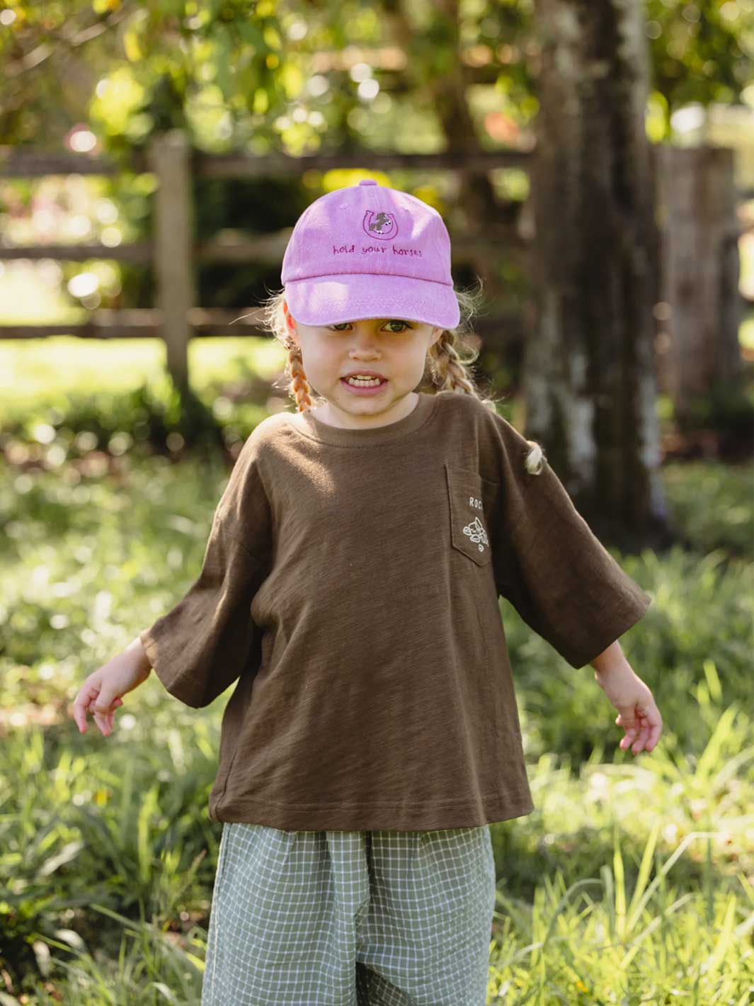 A young child standing outdoors in a grassy area, wearing a brown short-sleeve t-shirt, green and white plaid pants, and a pink baseball cap with embroidered text.
