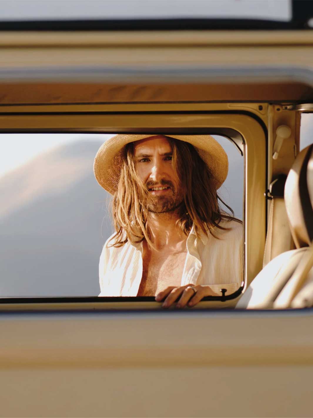 A man with long brown hair and a beard, wearing a straw hat and an open white shirt, looking out of a vintage beige vehicle window.