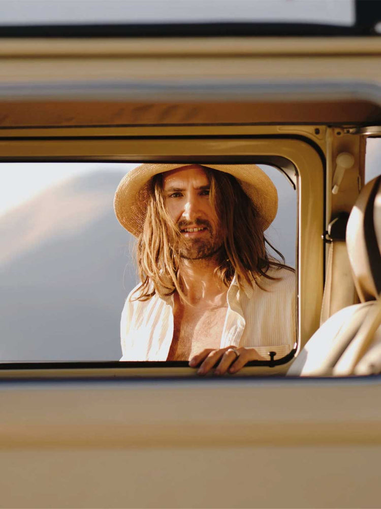 A man with long brown hair and a beard, wearing a straw hat and an open white shirt, looking out of a vintage beige vehicle window.