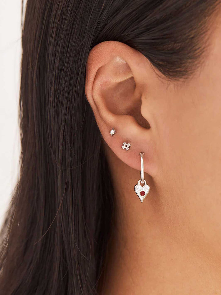 Close-up of an ear wearing a silver heart-shaped earring with a red gemstone.