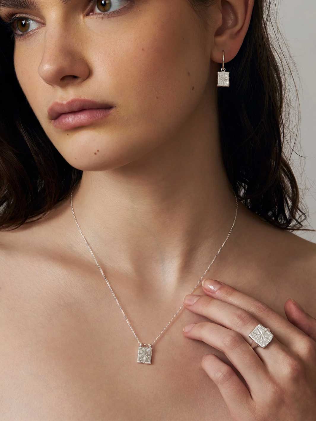 A close-up lifestyle shot of a model wearing a delicate silver chain necklace with a square pendant, matching silver hoop earring, and a silver ring with a square-cut stone.