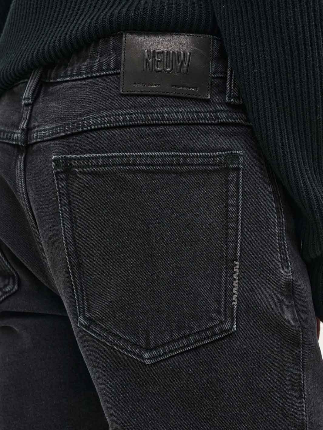 Close-up of the back waistband of black denim jeans with a black leather patch displaying the brand 'NEUW'. A black ribbed knit sweater is tucked into the waistband.
