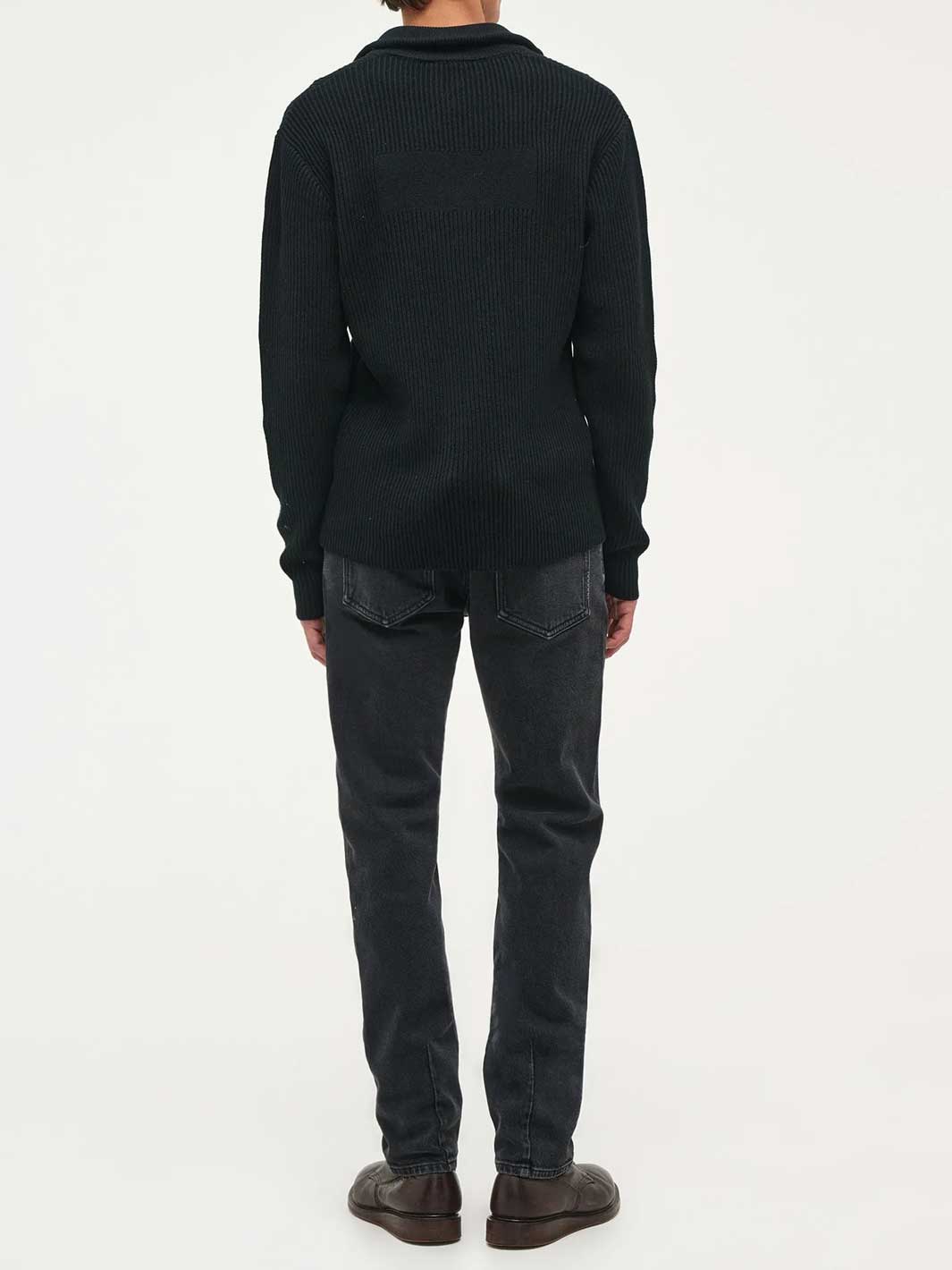 A person shown from the back wearing a dark ribbed knit sweater, dark black stone washed jeans, and brown shoes against a plain white studio background.