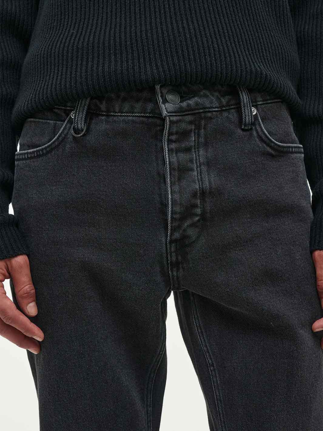 Close-up of a person wearing dark washed black denim jeans and a black ribbed sweater, showing the waistband, front pockets, and hands.