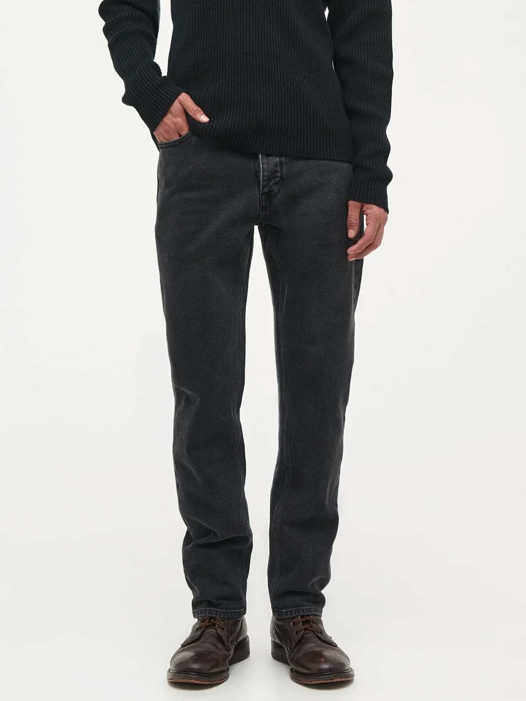 A person wearing slim-fit dark charcoal grey jeans, a black ribbed sweater, and dark brown leather shoes, standing against a plain white studio background.