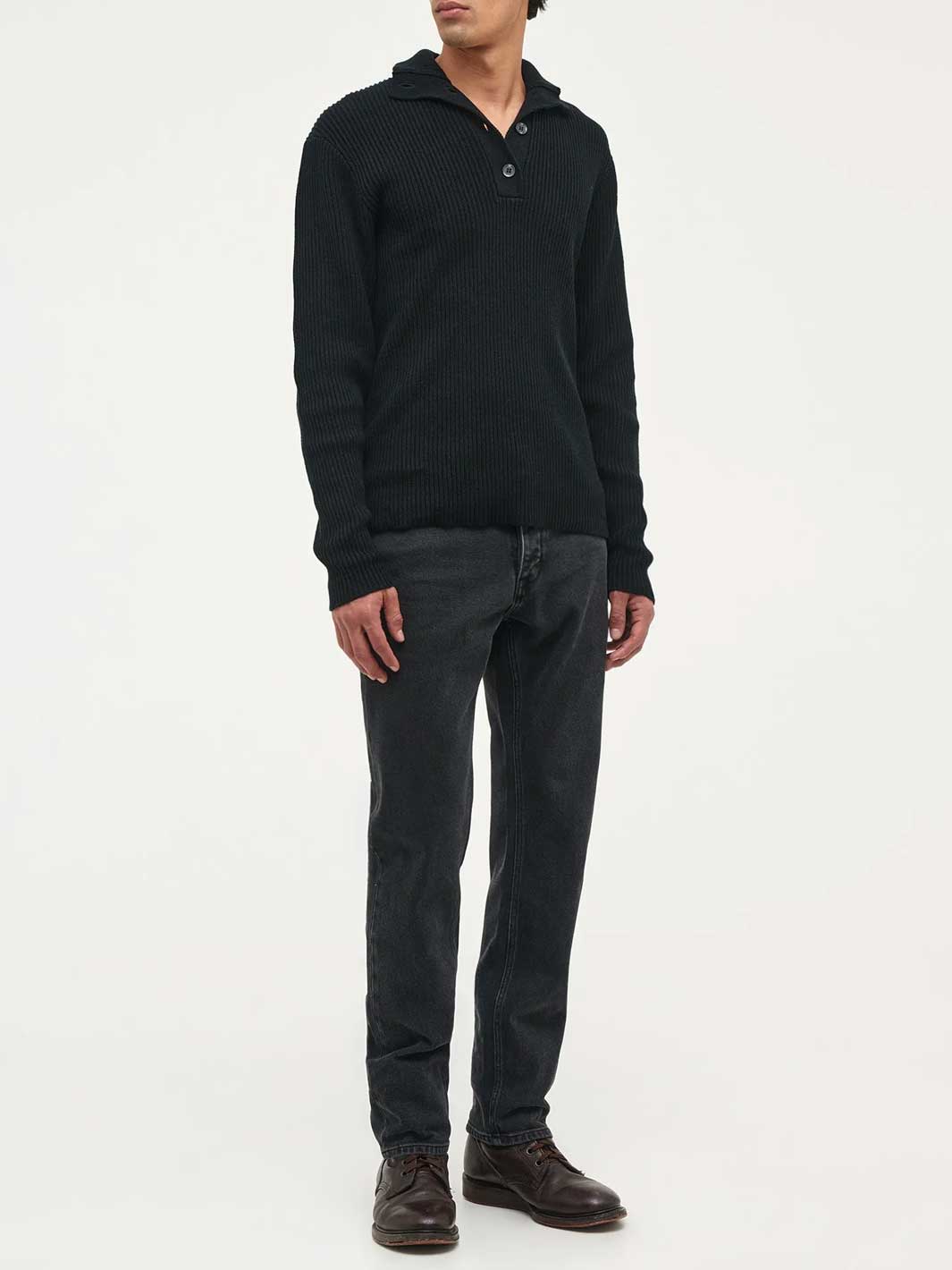 A man wearing a black ribbed knit polo-neck sweater, dark grey jeans, and brown leather shoes, standing against a plain white studio background.