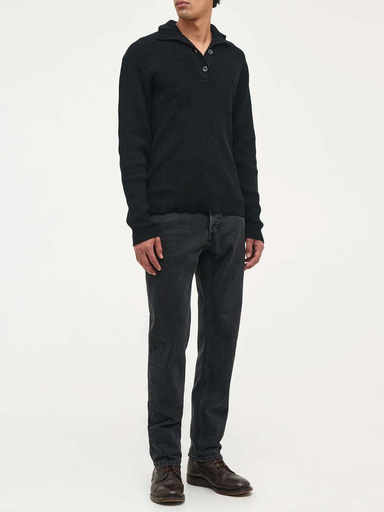 A man wearing a black ribbed knit polo-neck sweater, dark grey jeans, and brown leather shoes, standing against a plain white studio background.