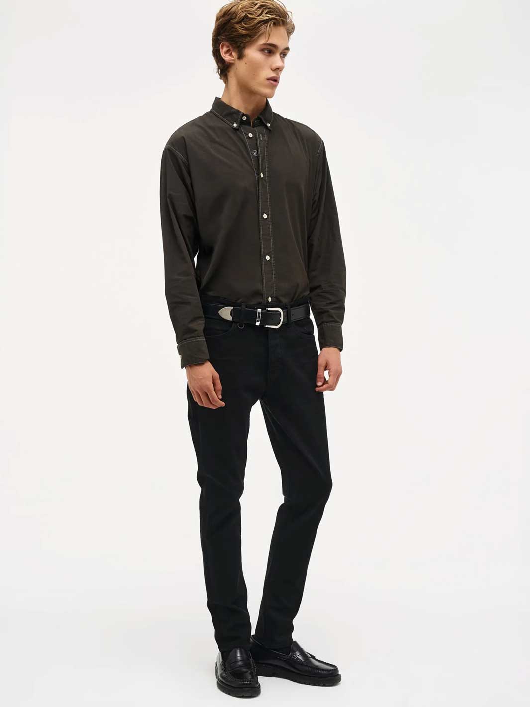 A man modelling a black long-sleeve button-down shirt, black denim jeans, a black belt, and black leather shoes against a plain white studio background.