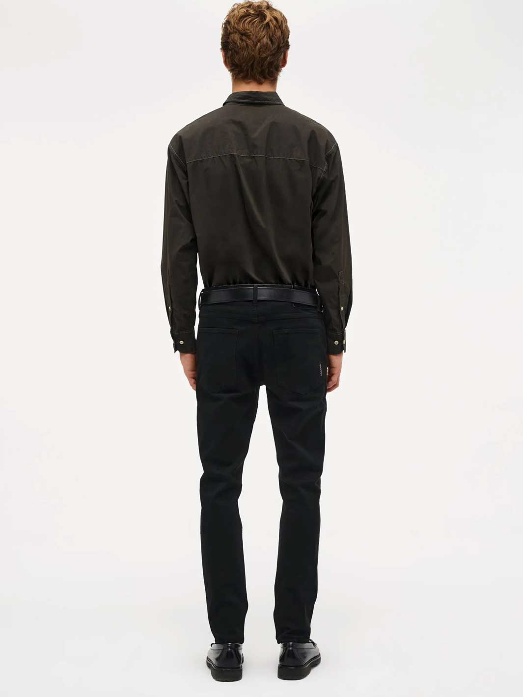A man viewed from behind wearing a dark charcoal long-sleeve button-up shirt, black trousers, and black leather shoes, standing against a plain white studio background.