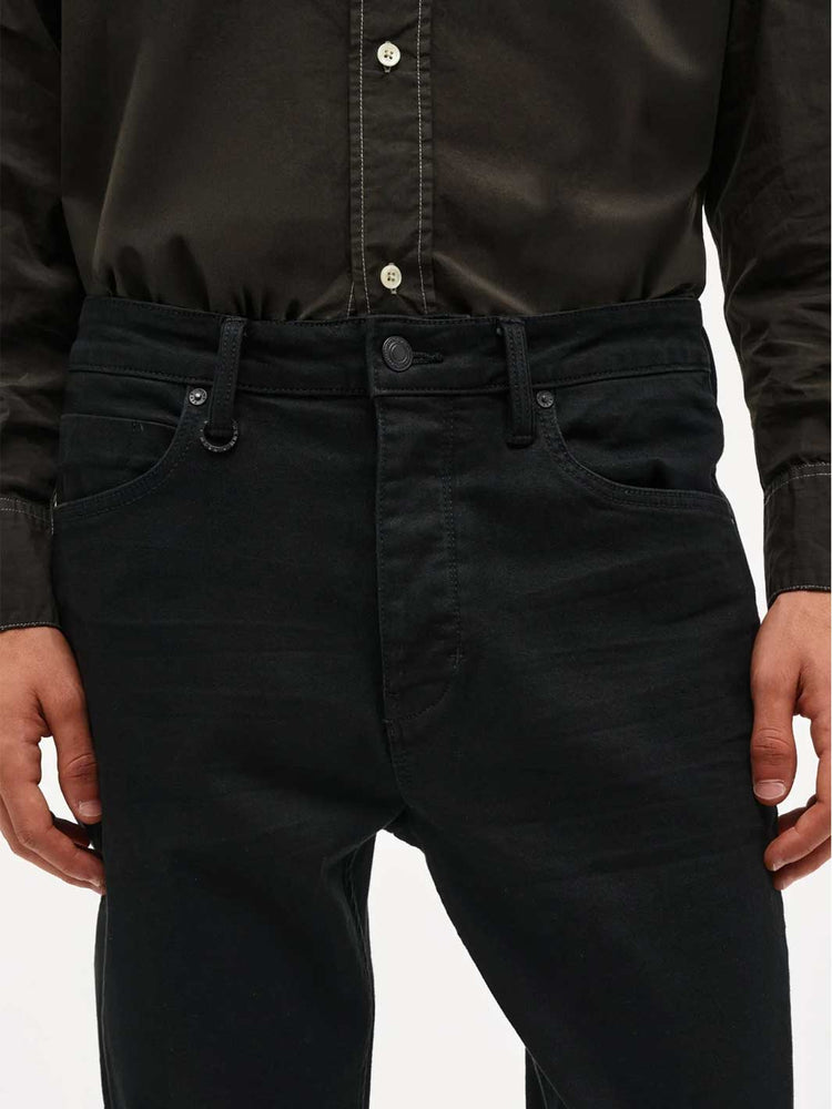 Close-up of a person wearing dark washed denim jeans and a black button-up shirt, showing the front waistband, belt loops, pockets, and hands against a plain light background.