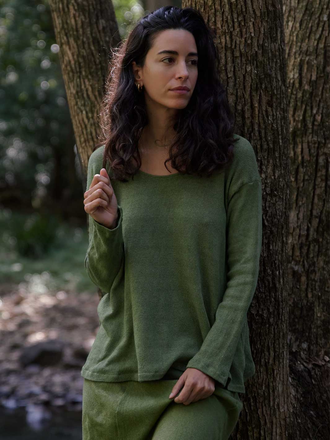 A woman with long dark curly hair stands outdoors leaning against a large tree trunk, wearing a matching olive green long-sleeve top and skirt set.