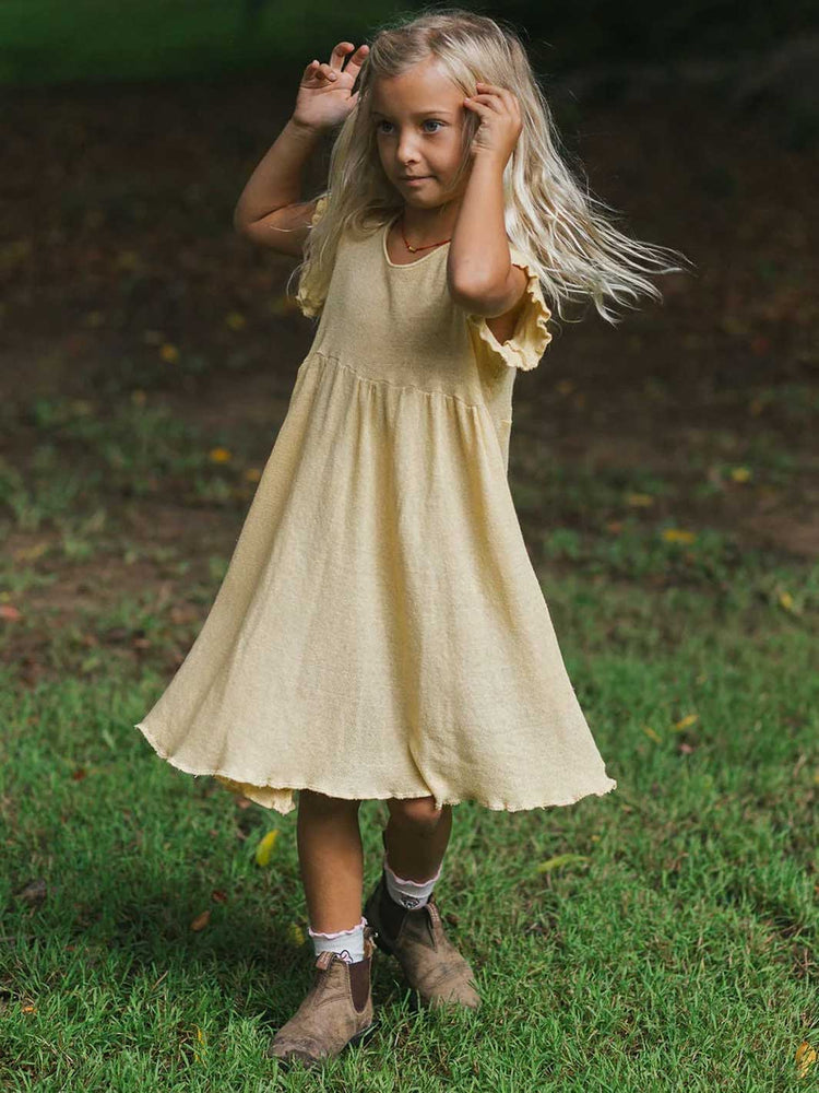 A young girl with long blonde hair stands on a grassy outdoor background wearing a light yellow cotton knit dress with ruffled sleeves and brown boots.