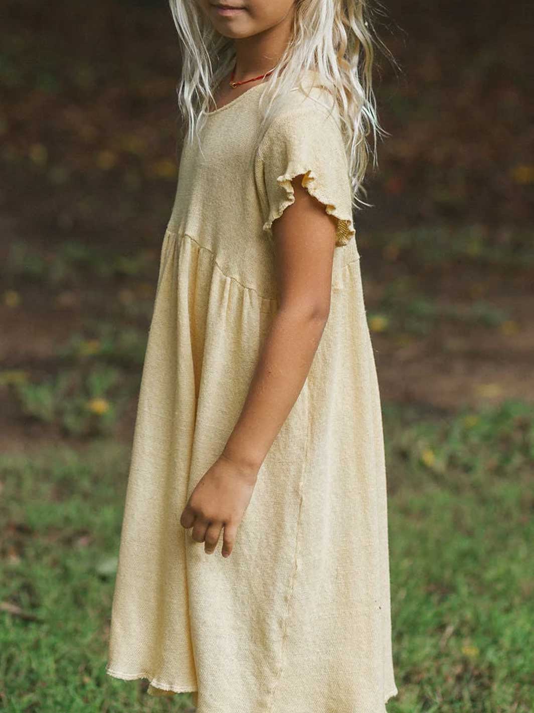 A young girl wearing a light yellow cotton knit dress with ruffled short sleeves, standing outdoors in a grassy, wooded area.