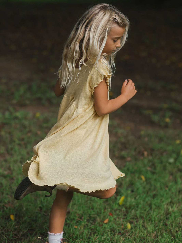 A young girl with long blonde hair wearing a pale yellow dress and dark boots, captured mid-stride while running outdoors on a grassy lawn.