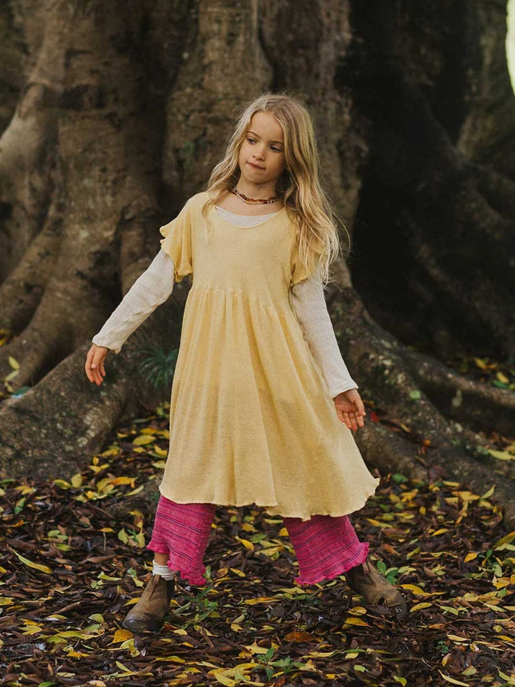 A young girl stands outdoors in front of a large tree trunk, wearing a pale yellow tunic dress over a white long-sleeve shirt, bright pink leggings, and brown boots, with fallen leaves on the ground.