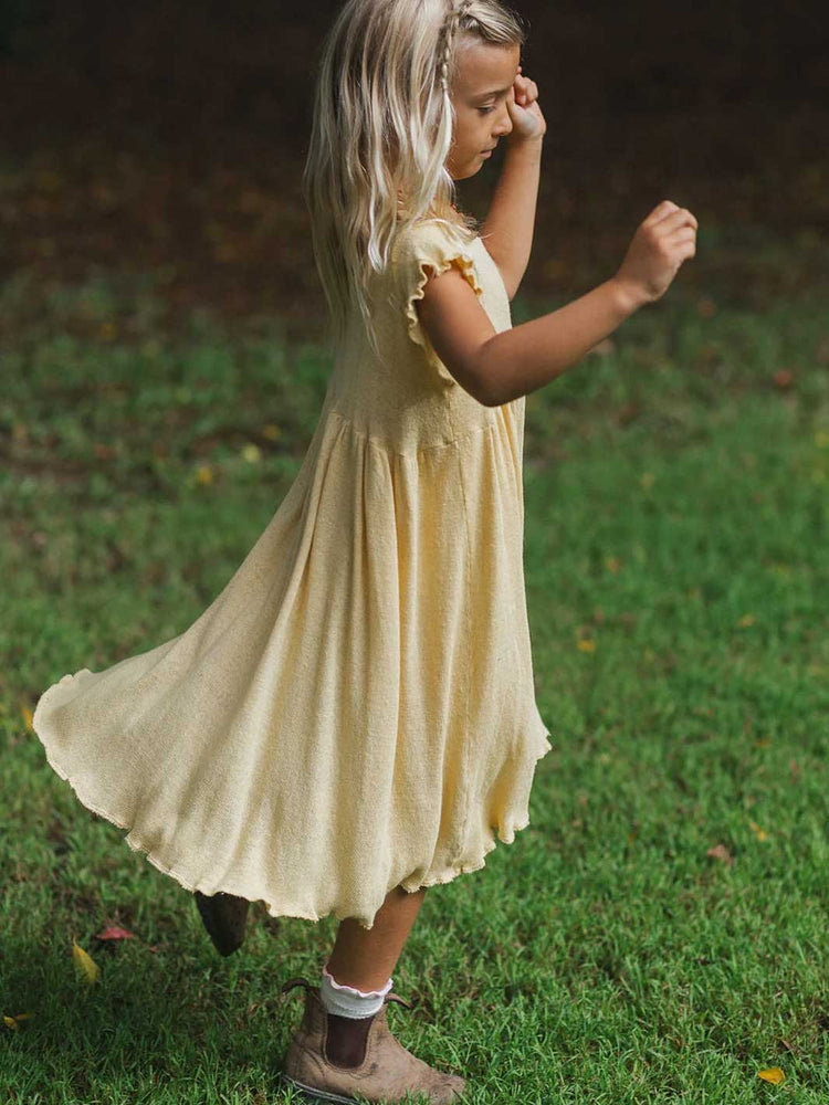 A young girl with long blonde hair wearing a flowing pale yellow dress, standing on a green grassy lawn.