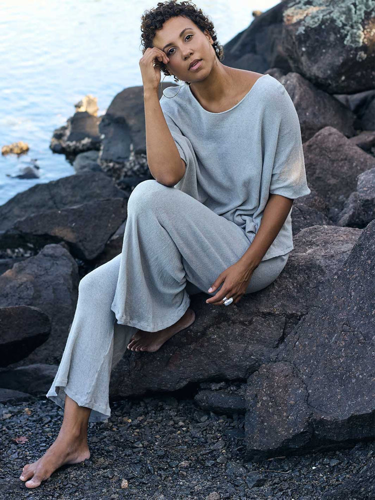 A woman sits barefoot on large dark rocks by the water, wearing a loose light gray linen-blend top and matching wide-leg pants.