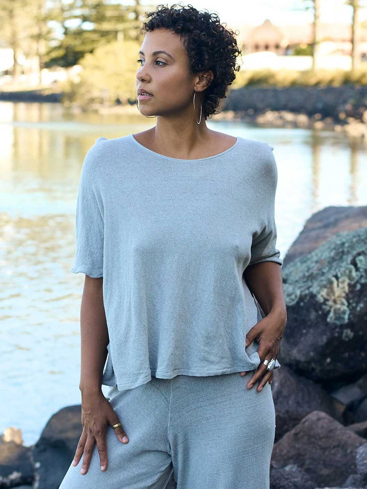 A woman wearing a light gray short-sleeve top and matching gray pants stands outdoors near water and rocks, looking off-camera.